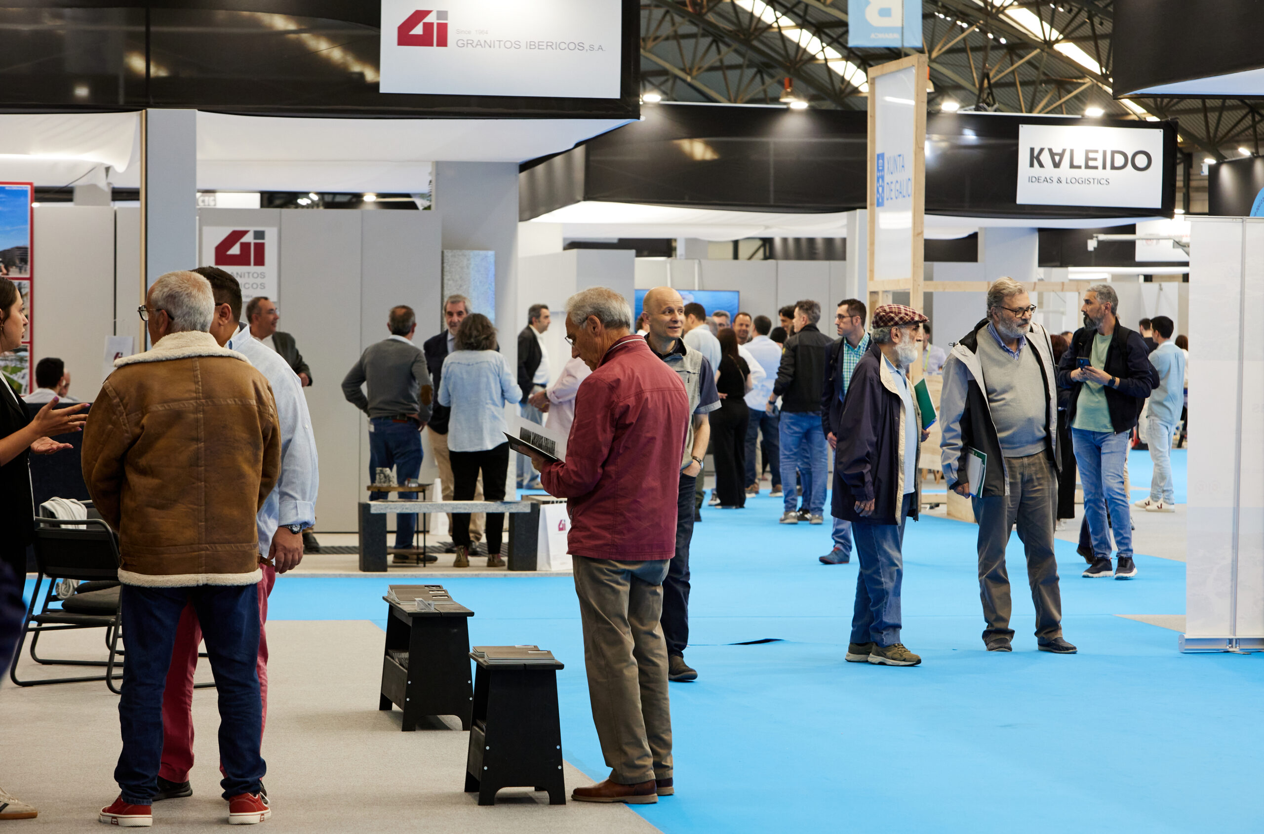 Imagen en una feria comercial donde se observa un espacio interior con varios asistentes conversando y mirando stands. Destacan logos visibles de marcas, principalmente Granitos Ibéricos y Kaleido, en un entorno profesional de exposición.