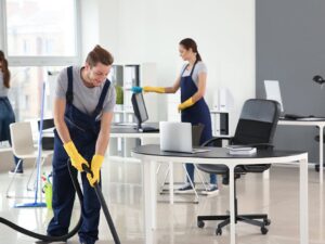 Fotografía de un grupo de personas en uniforme realizando labores de limpieza en una oficina moderna con grandes ventanas y mobiliario blanco y negro, transmitiendo eficiencia y profesionalidad en el servicio de limpieza.